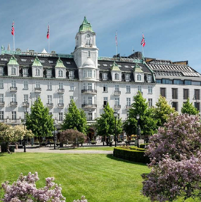 Façade of Grand Hotel Oslo by Scandic on sunny day. Four Norwegian flags on top of building and green trees in front.